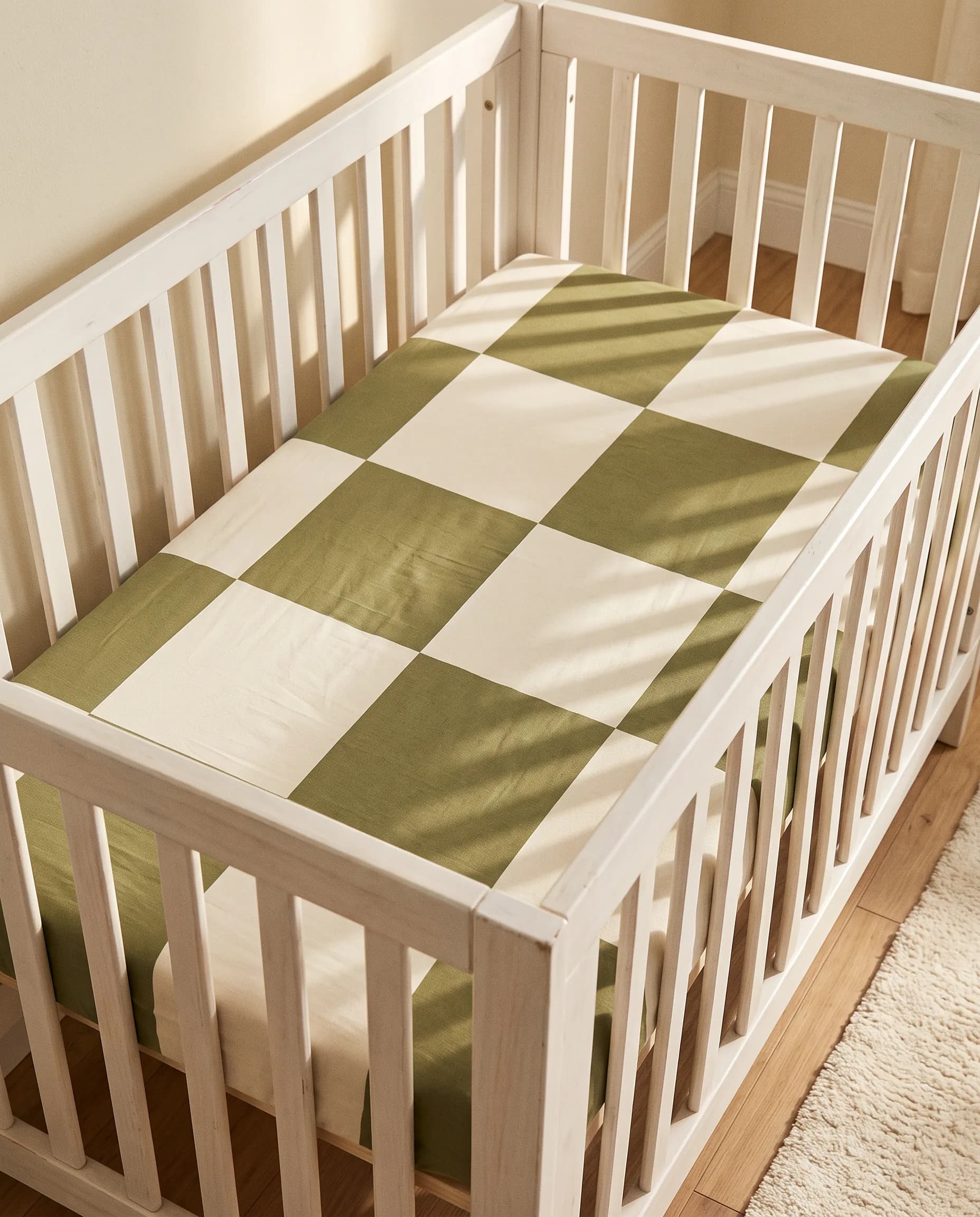 A white wooden crib fitted with the Olive Grove checker crib sheet in soft afternoon light
