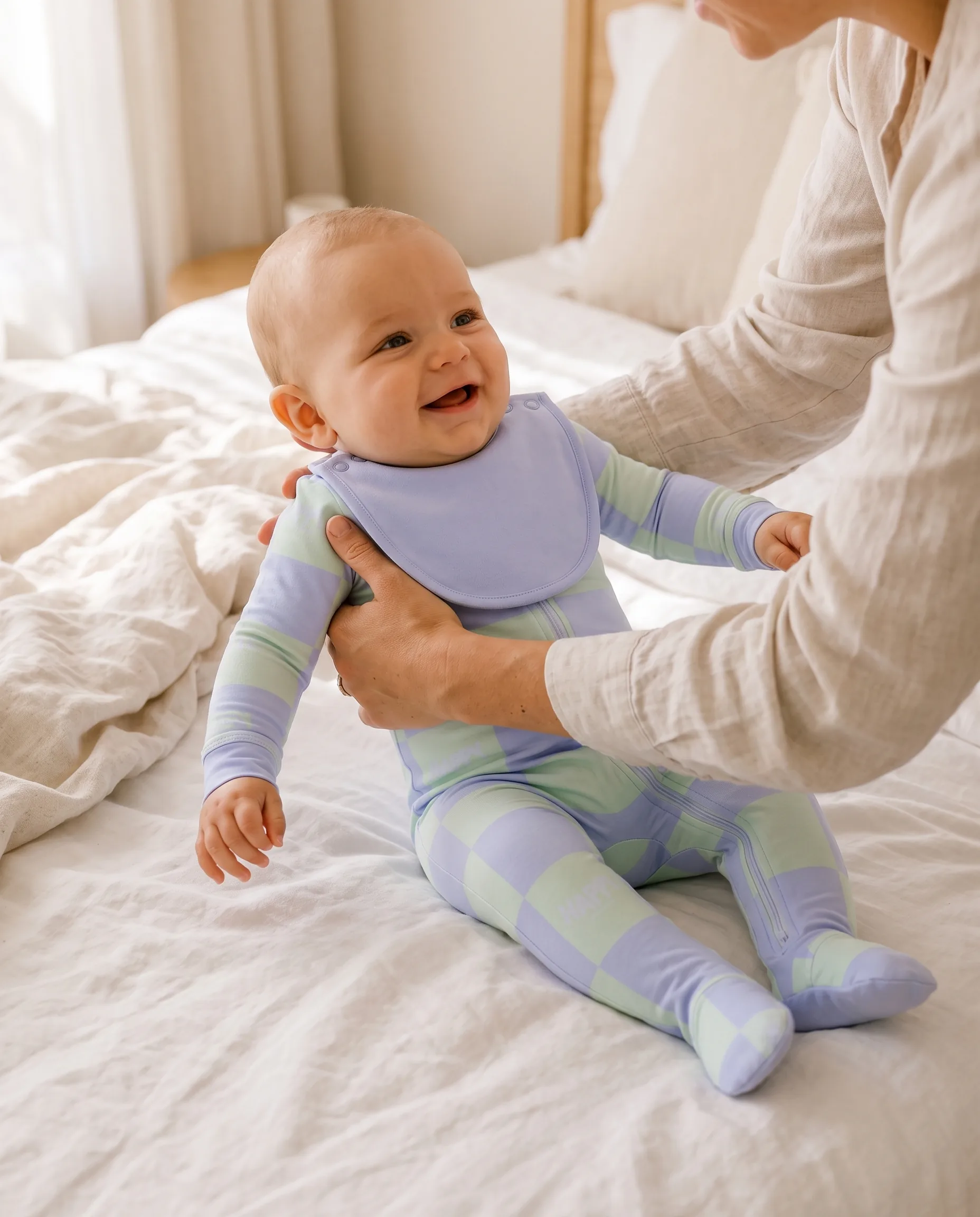 A parent holding a smiling baby on a white bed, the baby in the Dream Garden Everything Zippy and bib